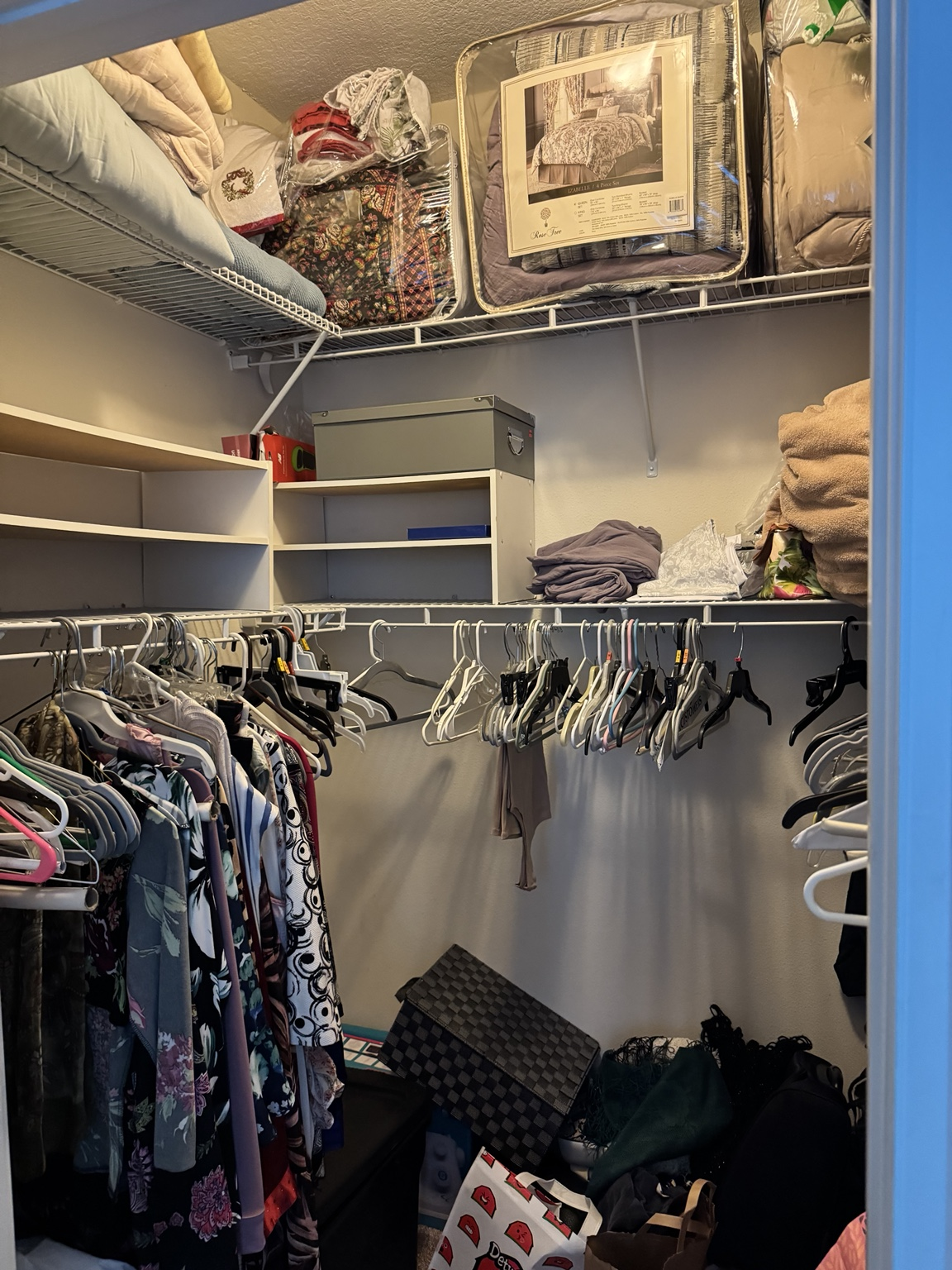Bedroom walk-in closet full of hanging clothing, bedding sets and stored boxes on upper shelves